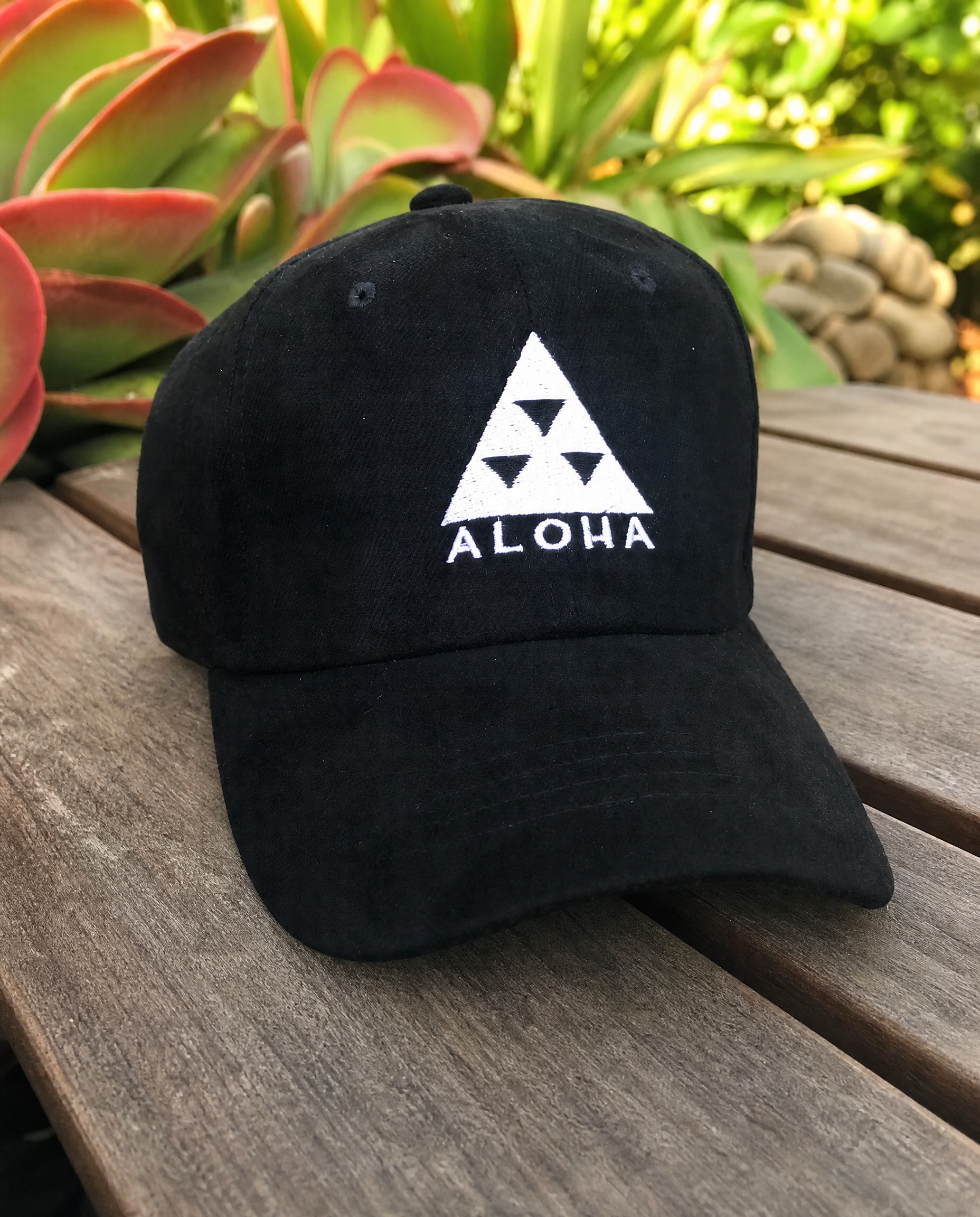 Black Aloha logo baseball cap photographed on a tropical garden table